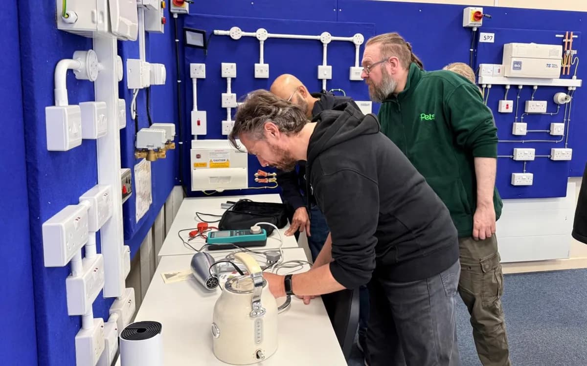Three trainees practice electrical wiring and testing on demonstration equipment in a blue-walled training room.